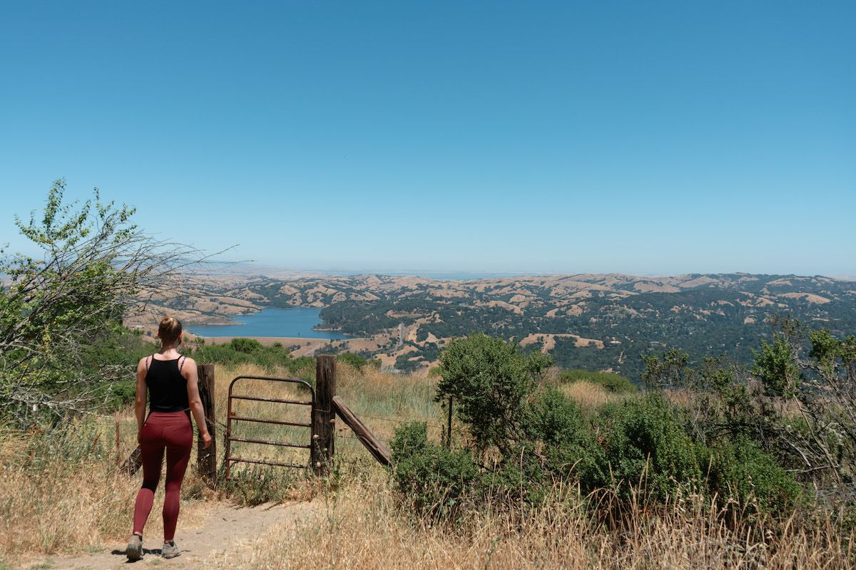 A woman wearing a black tank and dark red leggings walks toward an old gate in a California farmland landscape.