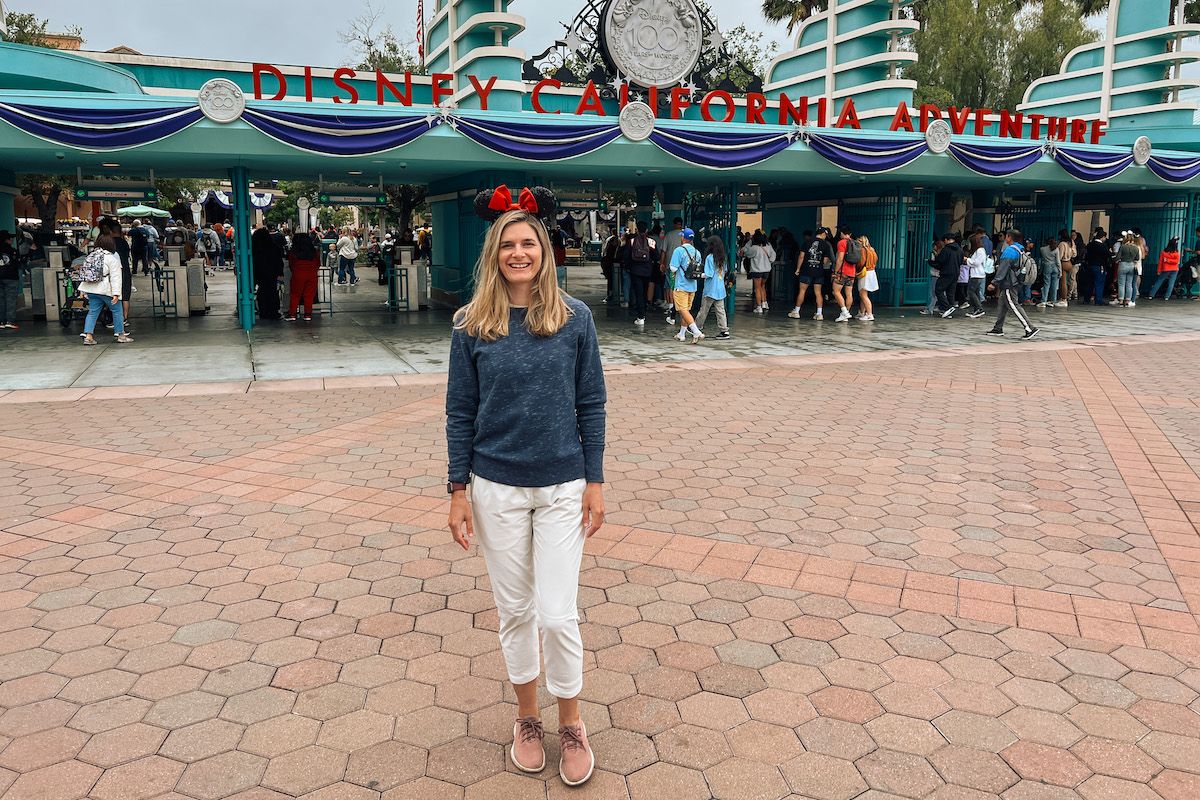 A young, light-haired woman in a blue sweater , white pants, pink Allbirds, and Minnie Mouse ears poses in front of the entrance to Disney California Adventure.