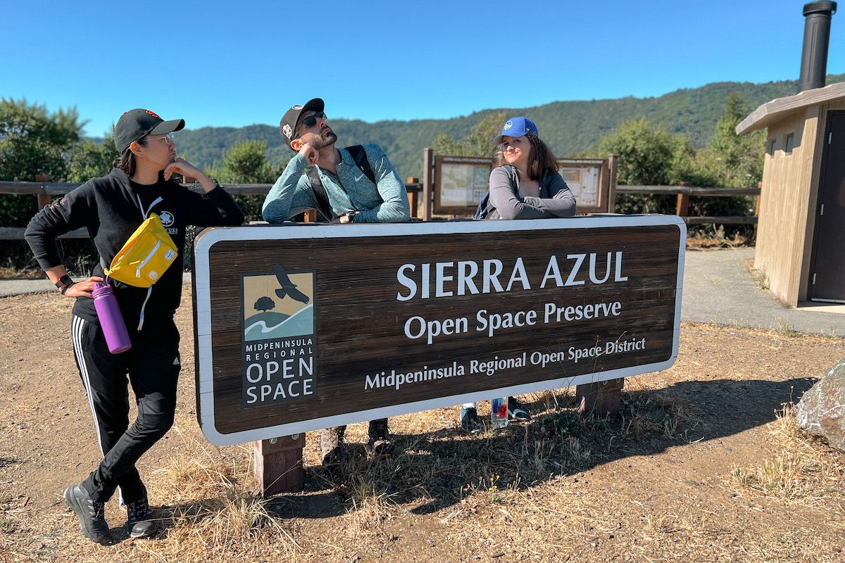 Three hikers wearng baseball caps pose leaning against a trailhead sign for the Sierra Azul Open Space Reserve, with tree-covers hills in the background behind them.