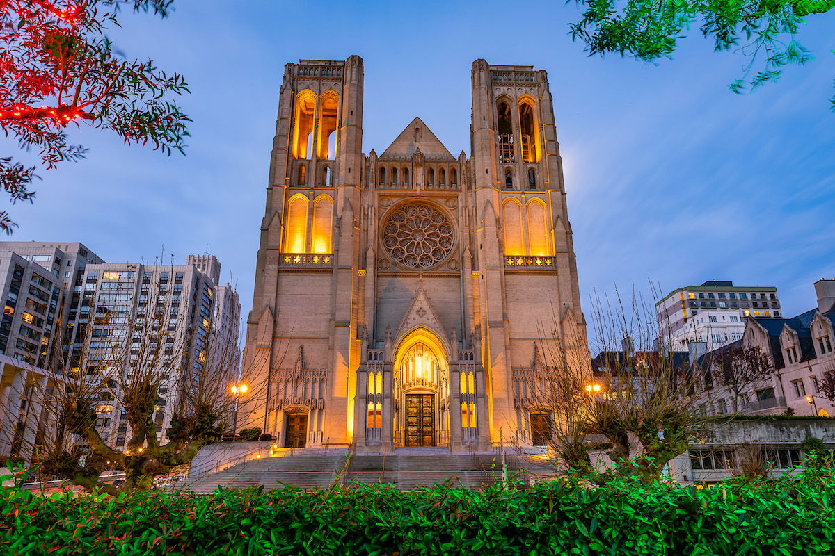 The facade of Grace Cathedral light up with a warm glow in fading, early evening light, with knobbly trees and bushes in the foreground.