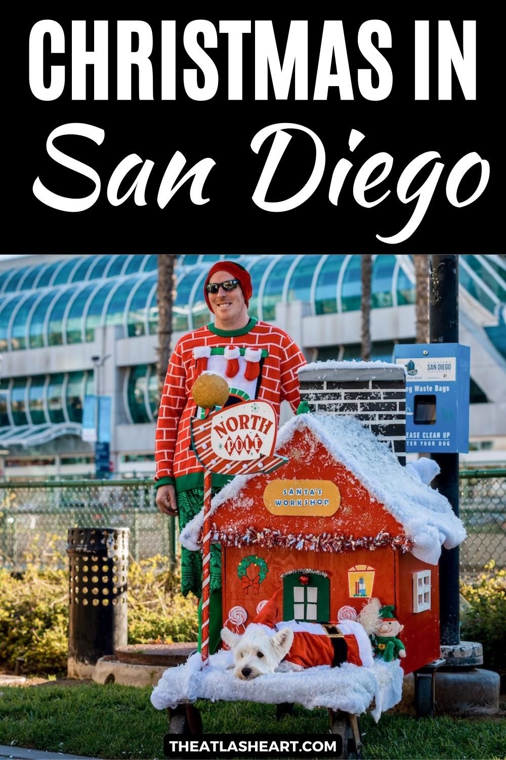 A man wearing an ugly Christmas sweater stands next to a small doghouse decorated like Santa's workshop, with a small, white dog in a Santa costume lying out front, and palm trees in the background, with the text overlay, "Christmas in San Diego."