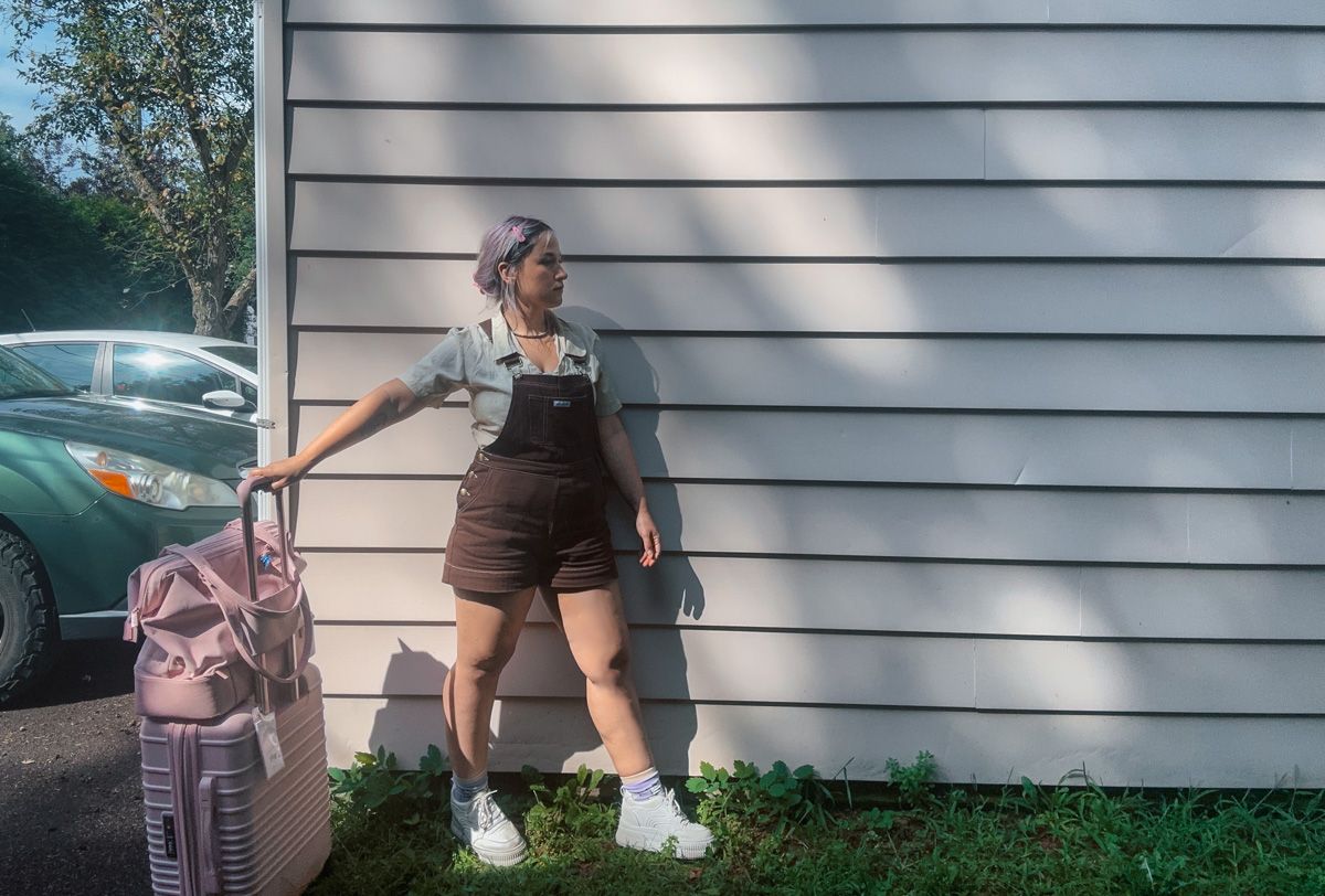 A woman with purple hair wearing brown overall shorts stands in front a the side of a light beige wooden house, with her hand on a pink Beis suitcase, with a Beis Mini Weekender bag on top of it.