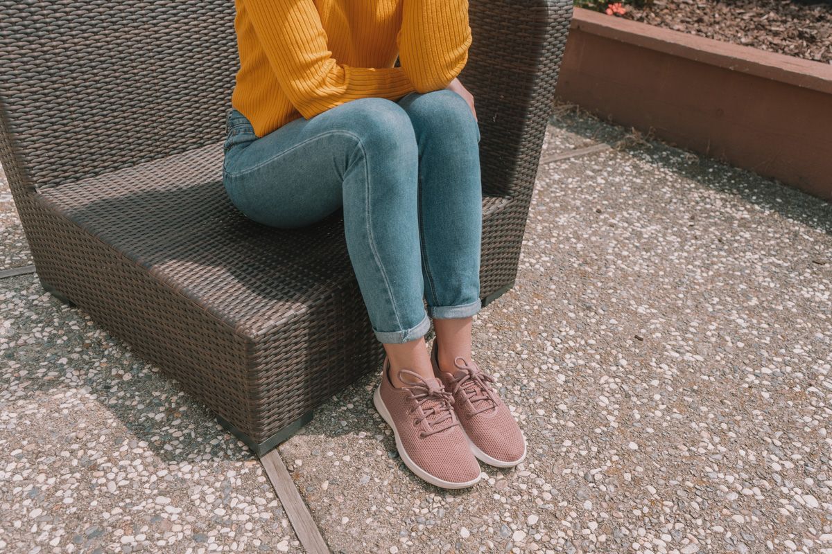 The lower half of a woman wearing a yellow sweater, jeans, and pink Allbirds sitting on a wicker patio chair.