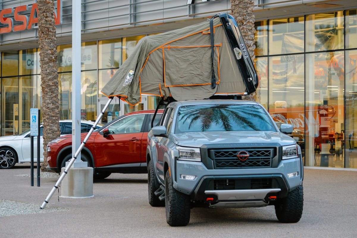 A grey truck with a taupe truck bed tent parked in a Nissan dealership parking lot with several cars in the background.