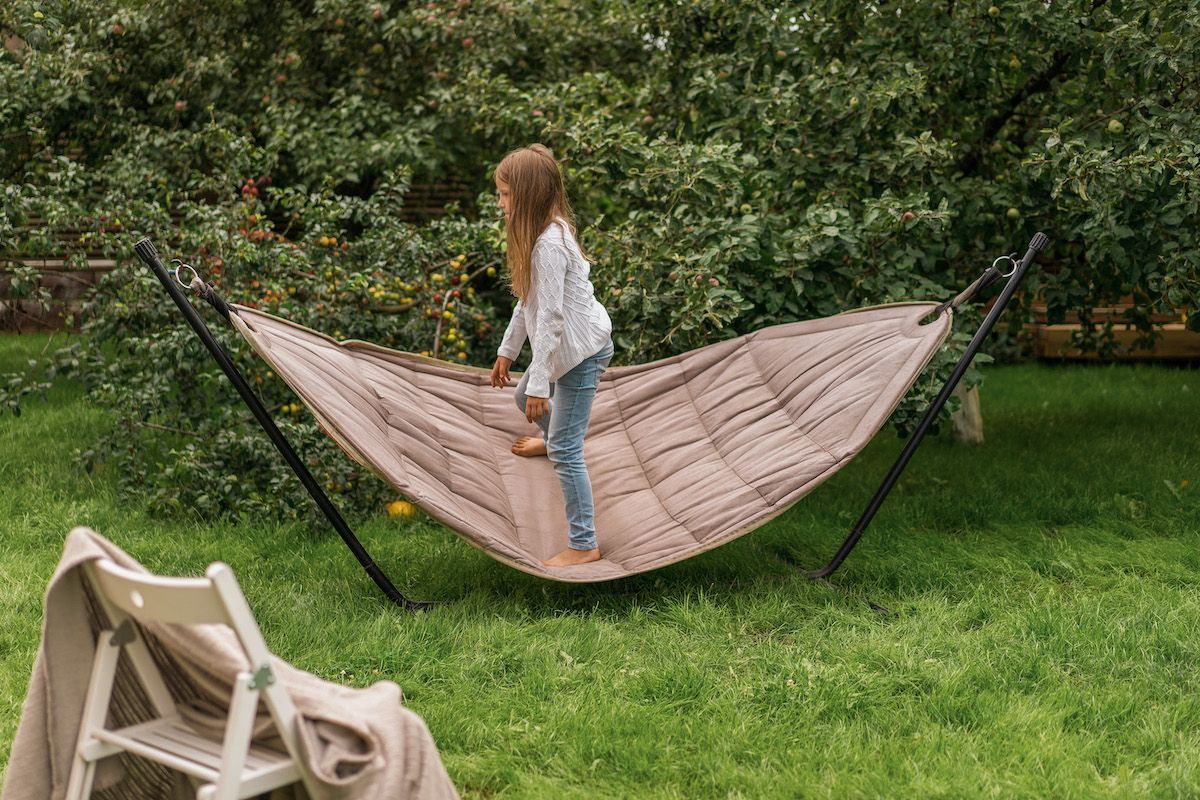 A young blonde girl wearing jeans and a white sweater stands in. aprofile on a grey hammock supported by a stand next to an apple tree in a backyard setting.