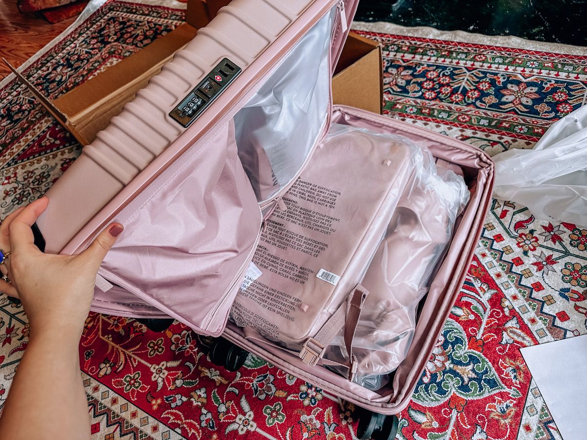 A hand opening a new, pink, Beis suitcase, with its packaging strewn across an oriental rug in the background. 