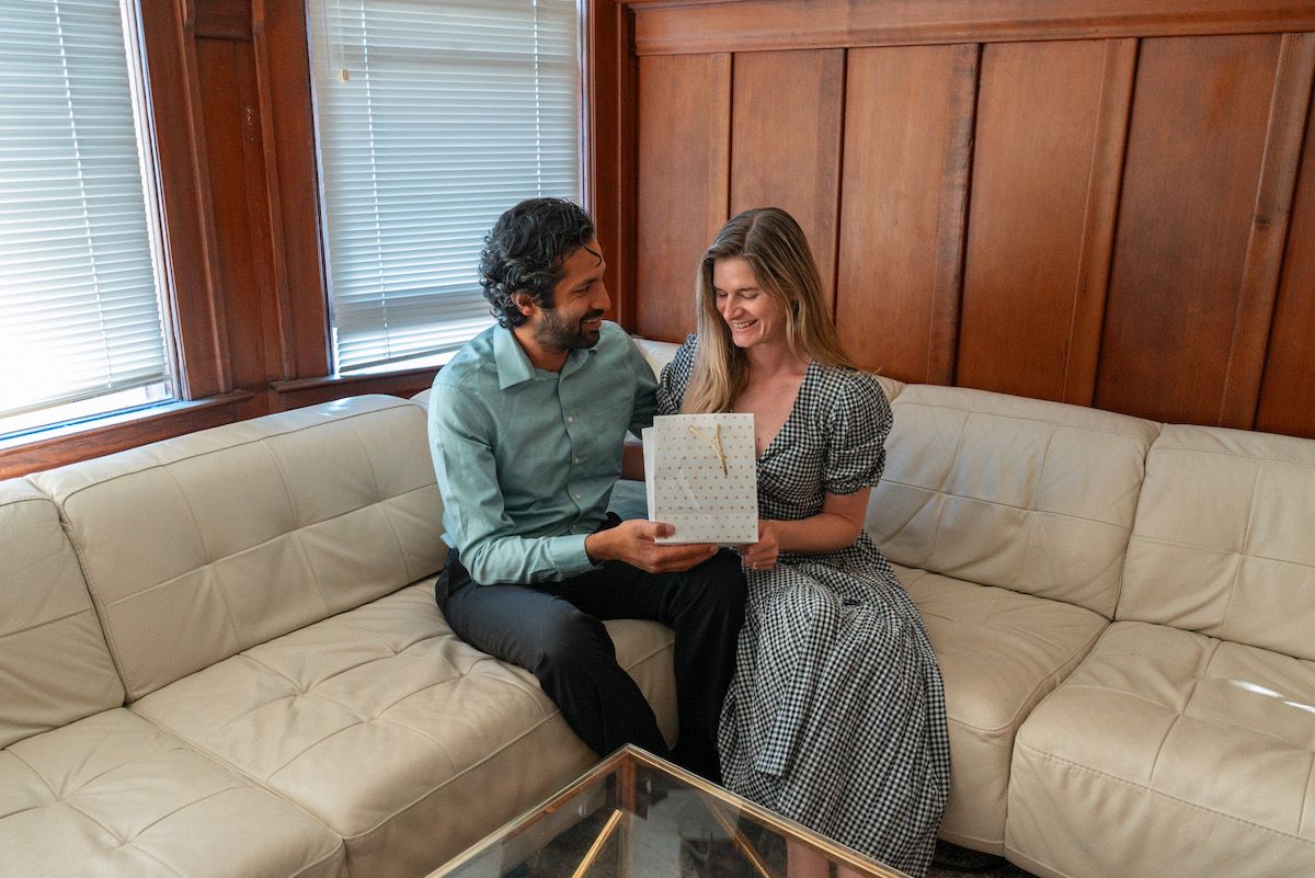 A man in a blue shirt affectionately gives a white gift bag to a woman in a gingham maxi dress as they sit in the corner of an L-shaped, white leather couch, with a wood-paneled interior in the background.