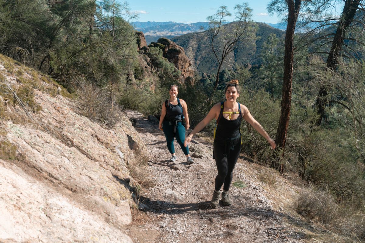 Two smiling women in leggings hiking up a rocky trail in a mountainous landscape.