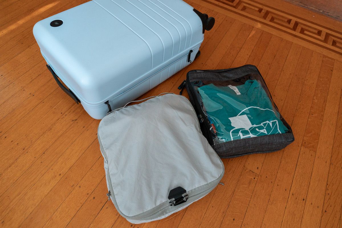 A light blue suitcase sits on a wooden floor with two packing cubes laid out on the floor beside it.