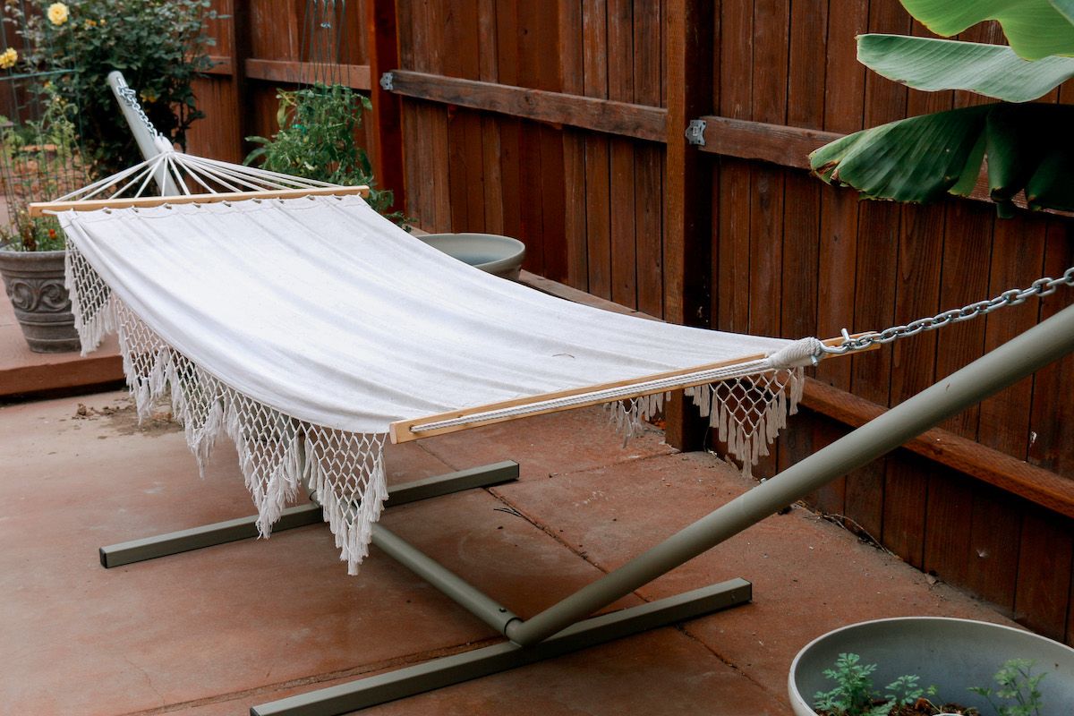 A white macrame hammock on a freestanding portable hammock stand next to a wooden fence on a patio in a backyard, banana leaves peaking in from the corner.