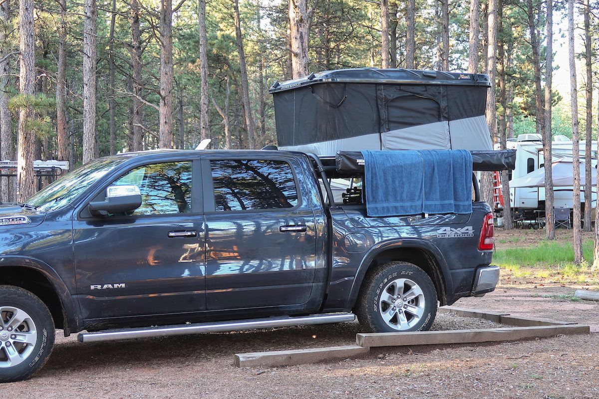 A dark grey truck with a black truck bed tent parked in a campsite with numerous pine trees in the background.
