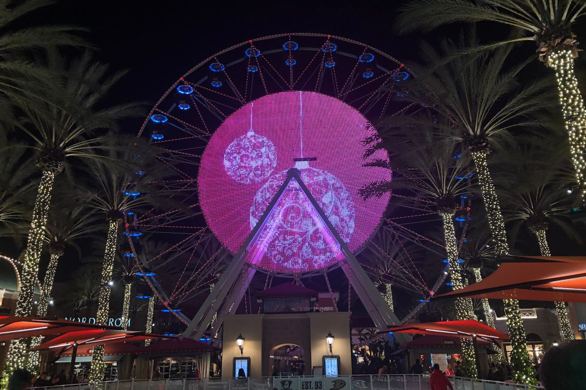 A ferris wheel seen at night sporting a  round, red, Christmas-themed light display on its side, and surrounded by palm trees decorated with Christmas lights.
