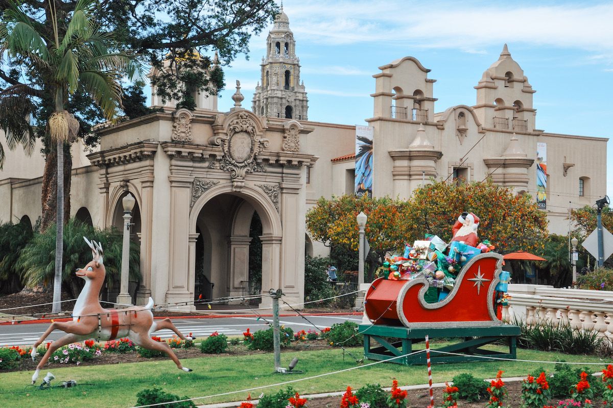 A display of Santa and his sleigh on the lawn in front of Spanish Mission-style building flanked by palm trees, with a partly cloudy blue sky behind it. 