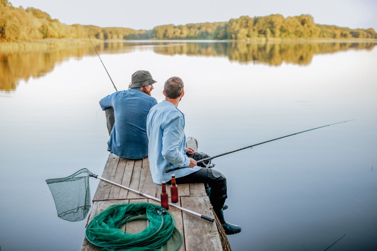 Two people in blue denim shirts seen from behind sitting on a dock on a lake, holding fishing poles and surrounded by fishing gear, which is among the best gifts for fishermen.