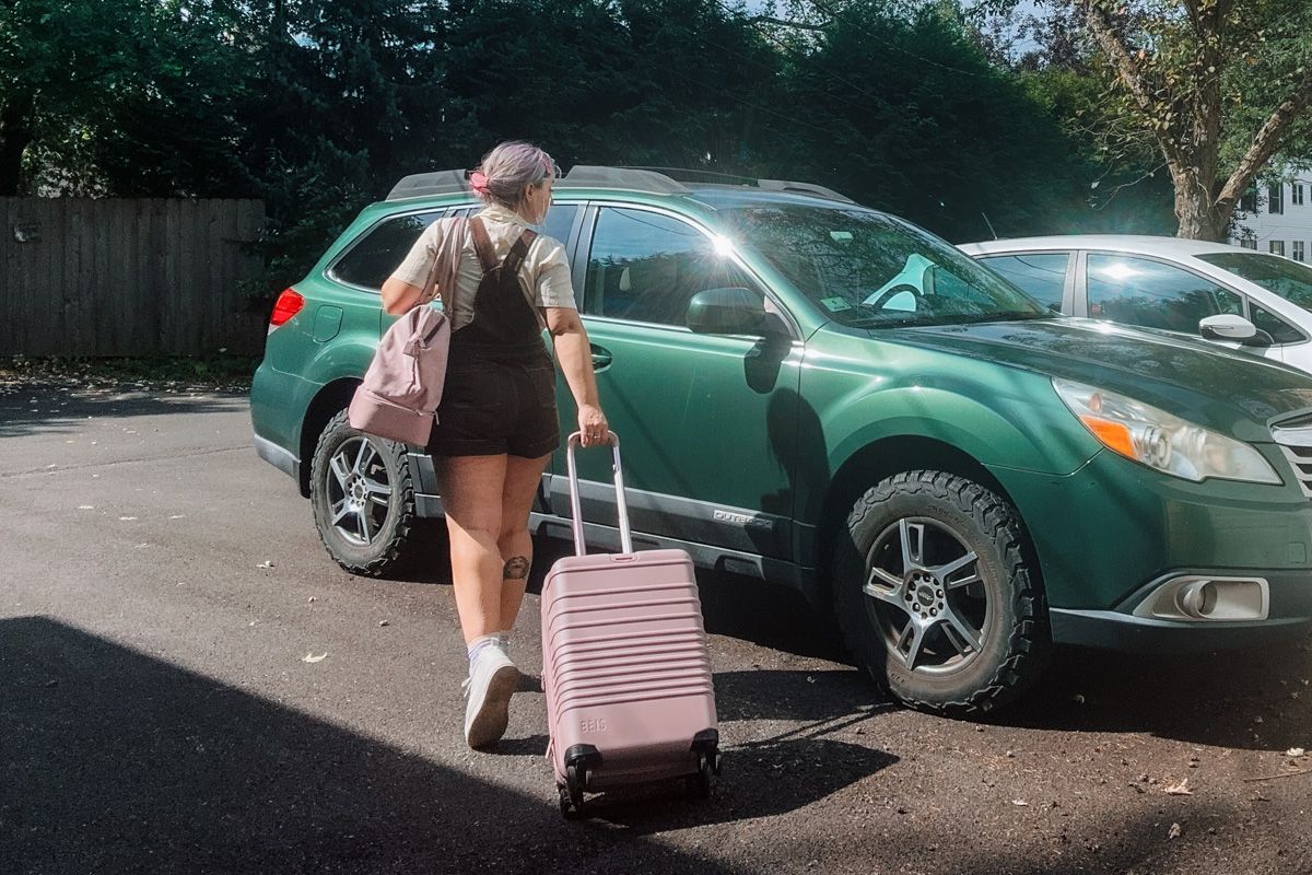 A woman in brown overall shorts seen from behind wheeling a pink suitcase and carrying a matching shoulder bag, walking past a green SUV in a parking lot, ready to write a Béis Luggage Review.