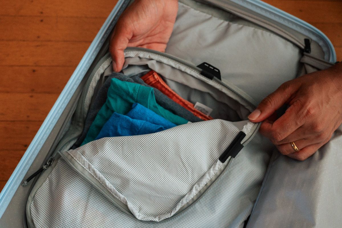 Are Packing Cubes Worth it? A pair of hands zipping up a light grey packing cube inside an open suitcase sitting on a hardwood floor.