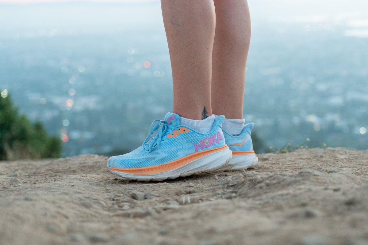 A close-up of a pair of feet wearing the Clifton 9 shoes, some of the best Hoka shoes for walking, standing on a rock at the top of a hill, with a foggy cityscape in the background.