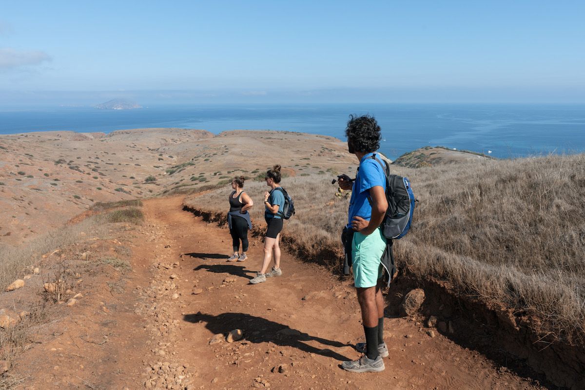 A male hiker in a blue shirt and aqua shorts looks down a red dirt hiking trail at two female hikers, with a clear blue sky and the ocean visible in the distance to show an example for this article, hiking for beginners.