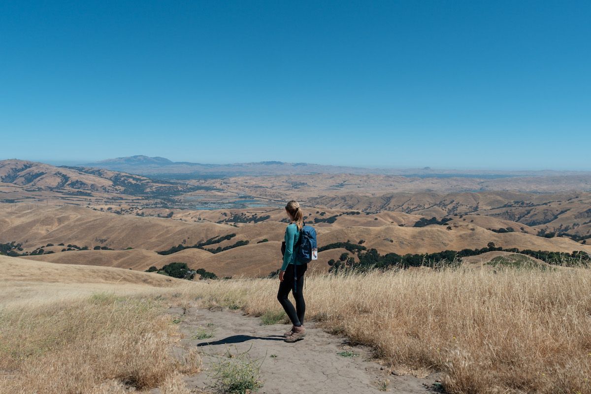 A woman in a blue backpack wearing an example of some of the best hiking leggings seen from behind as she walks down a dry grass-lined path in a California landscape of rolling golden hills.