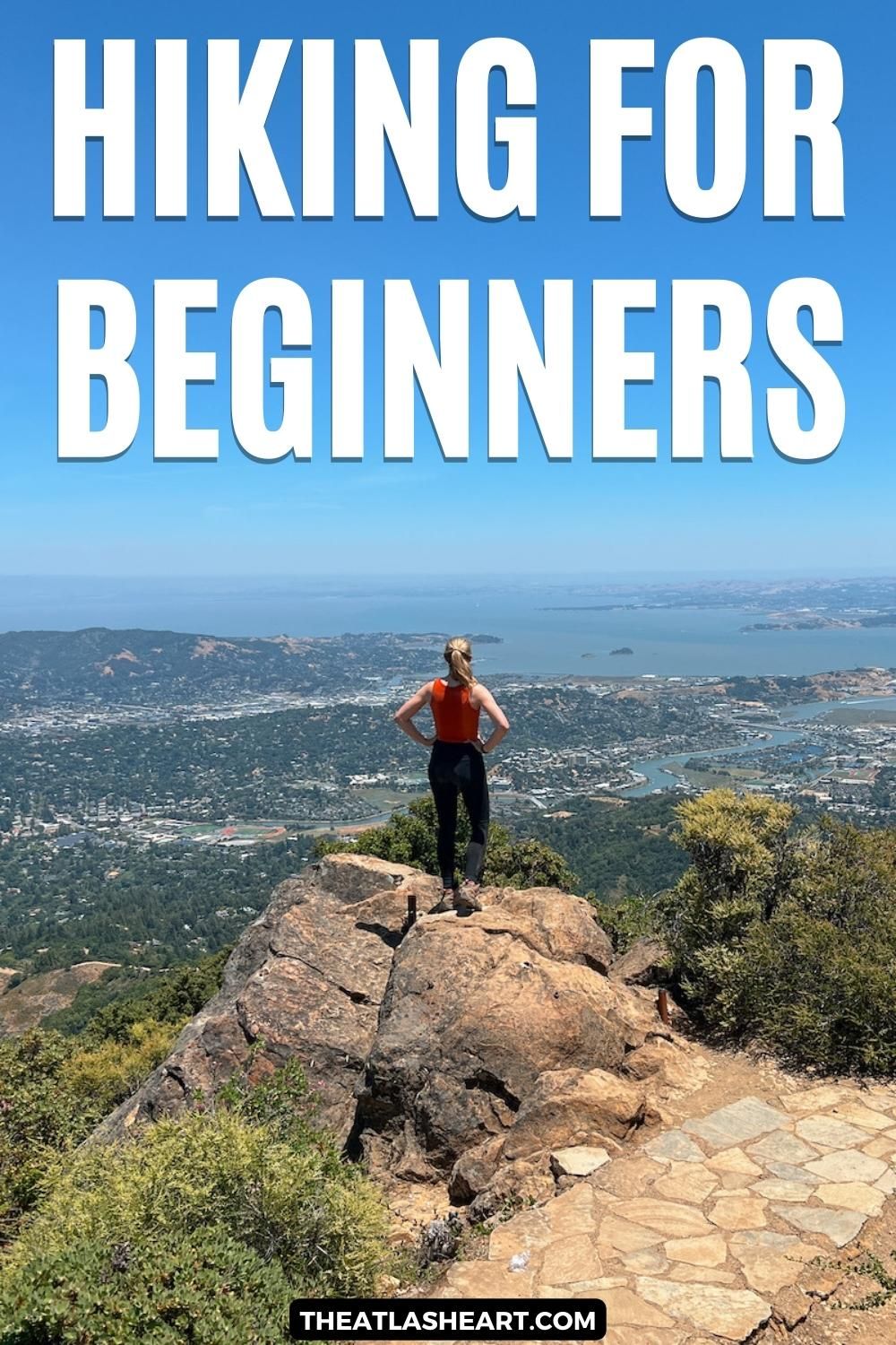 A woman wearing black leggings and an orange tank top seen from behind as she stands on a rocky overlook with a view of a cityscape and a bay beyond her, with the text overlay, "Hiking for Beginners."