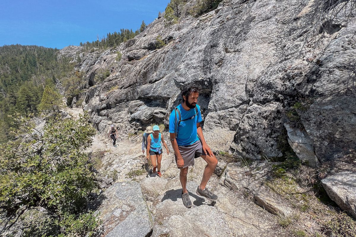 A man in a blue shirt hikes along a rocky, mountainous trail with a woman in a blue shirt close behind him, and two more people visible further down the path.