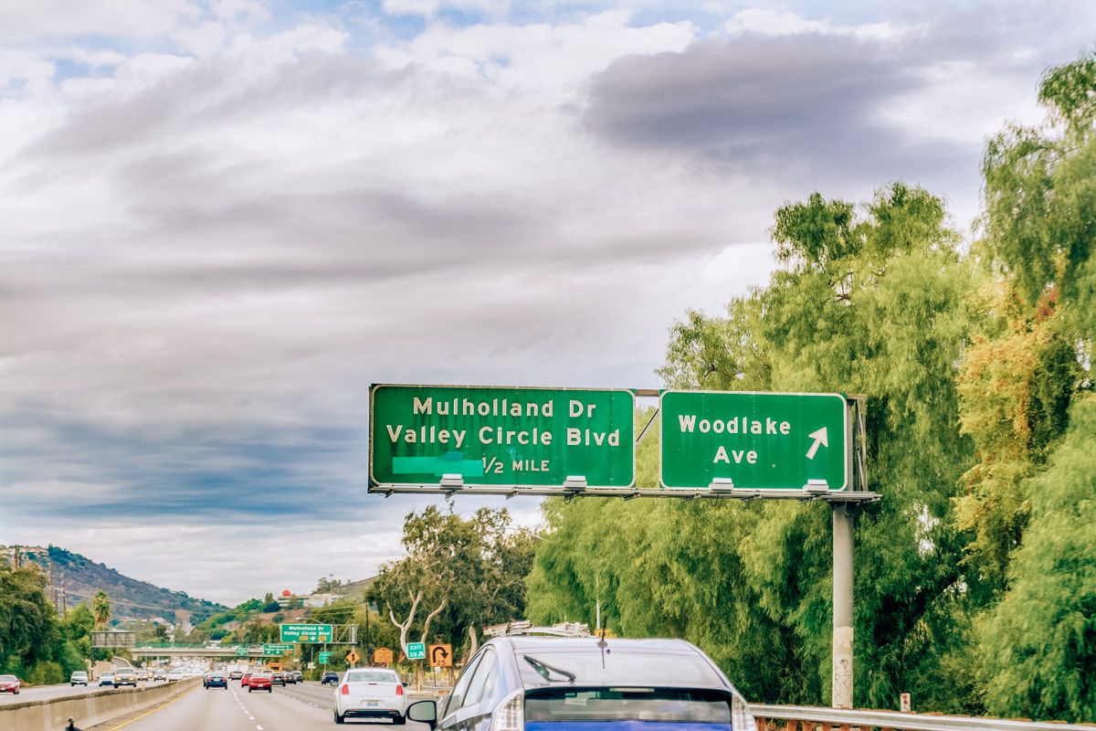 A scene on an LA freeway with two green signs, one reading "Mulholland Dr Valley Circle Blvd" and the other reading "Woodland Lake" with green leafy trees to the right and a partly cloudy sky overhead.