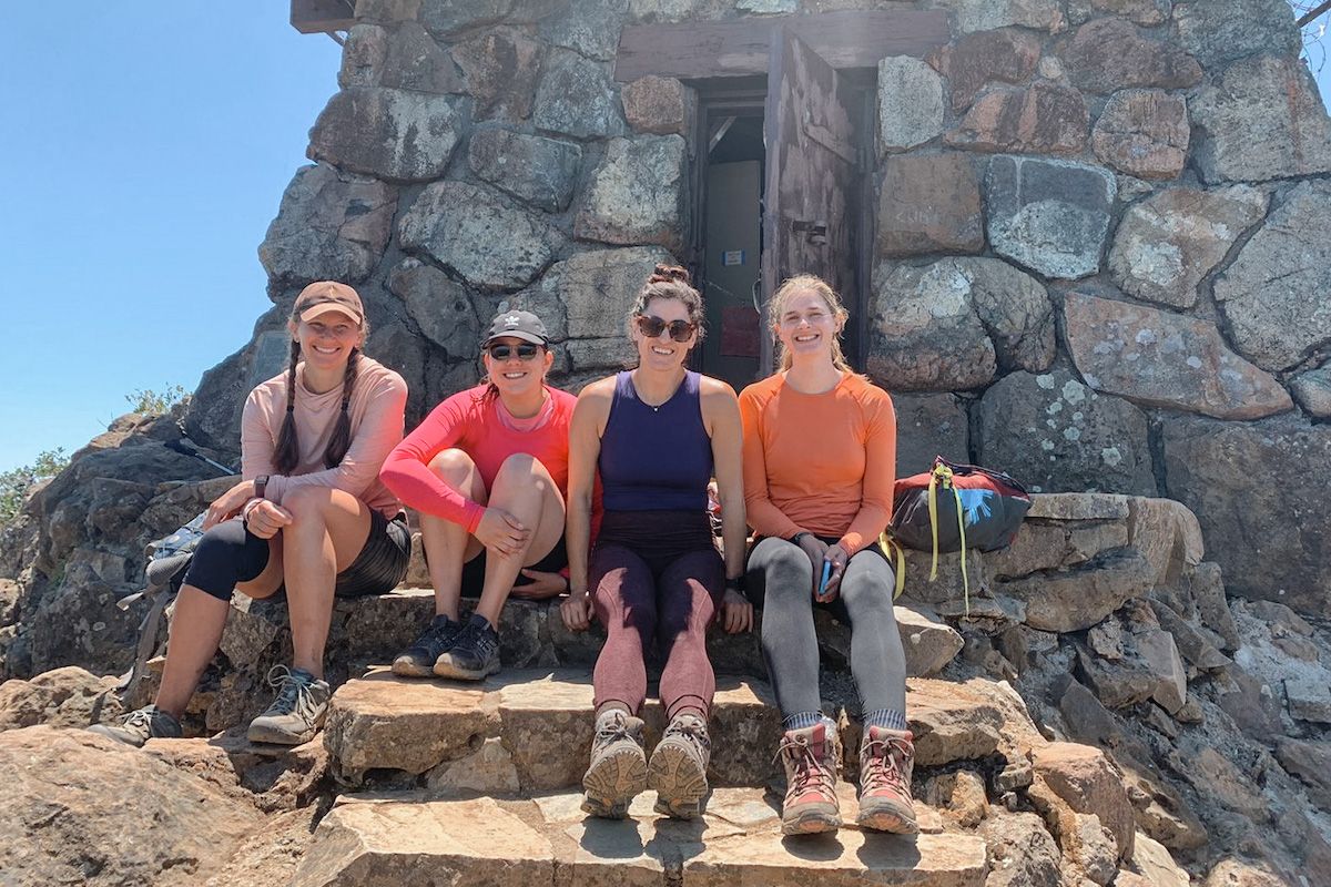 Four women in brightly colored hiking clothing sit in a line and smile at the camera on the steps of an old stone hut, with a clear blue sky behind it. 