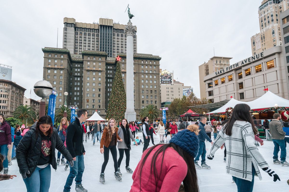 A crowd of people Ice Skating in Union Square in San Francisco, with Saks Fifth Avenue, a giant Christmas tree, and the Sir Francis Drake Hotel visible in the background.
