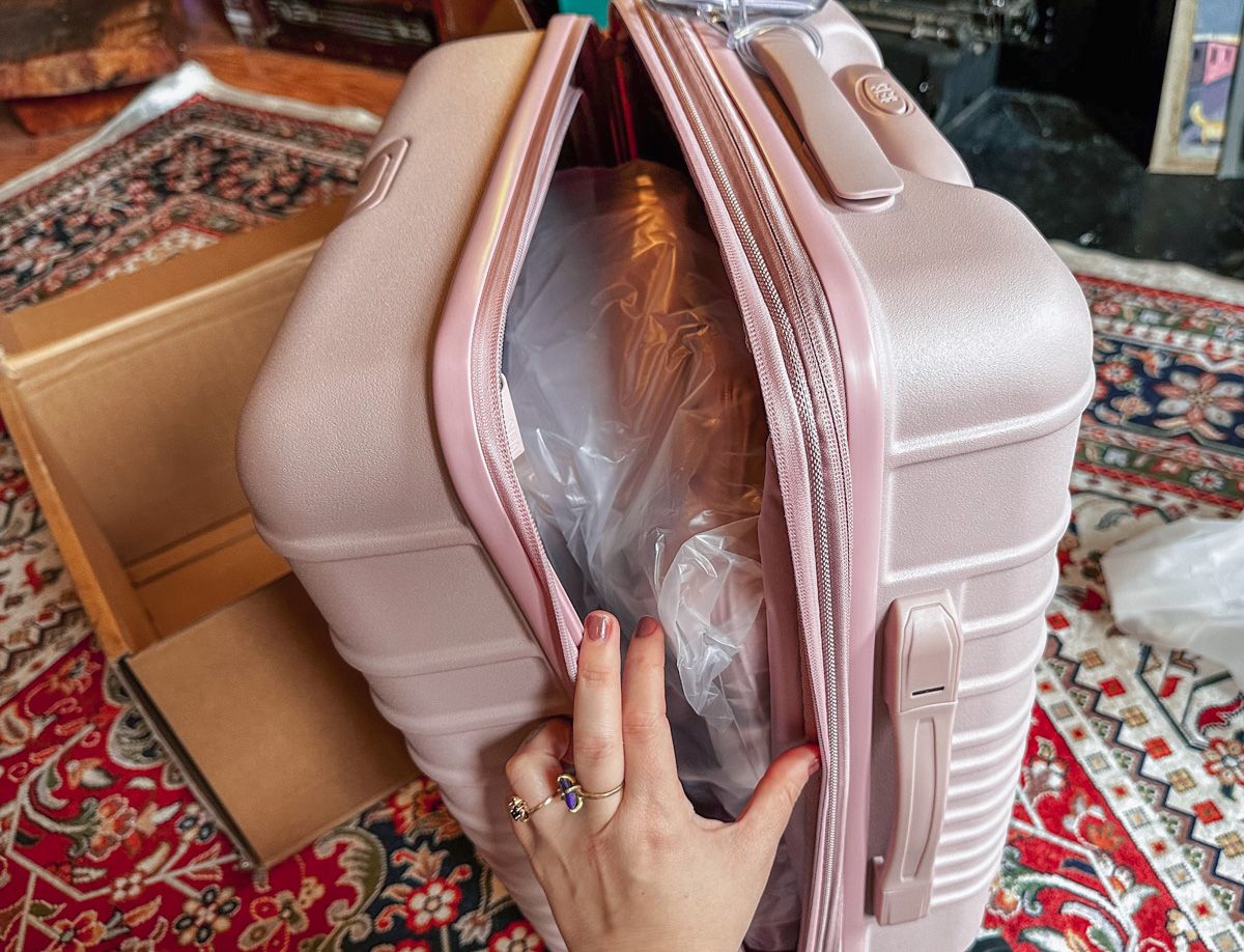 A hand opening the zipper of a new, pink, Beis suitcase, with its packaging strewn across an oriental rug in the background.