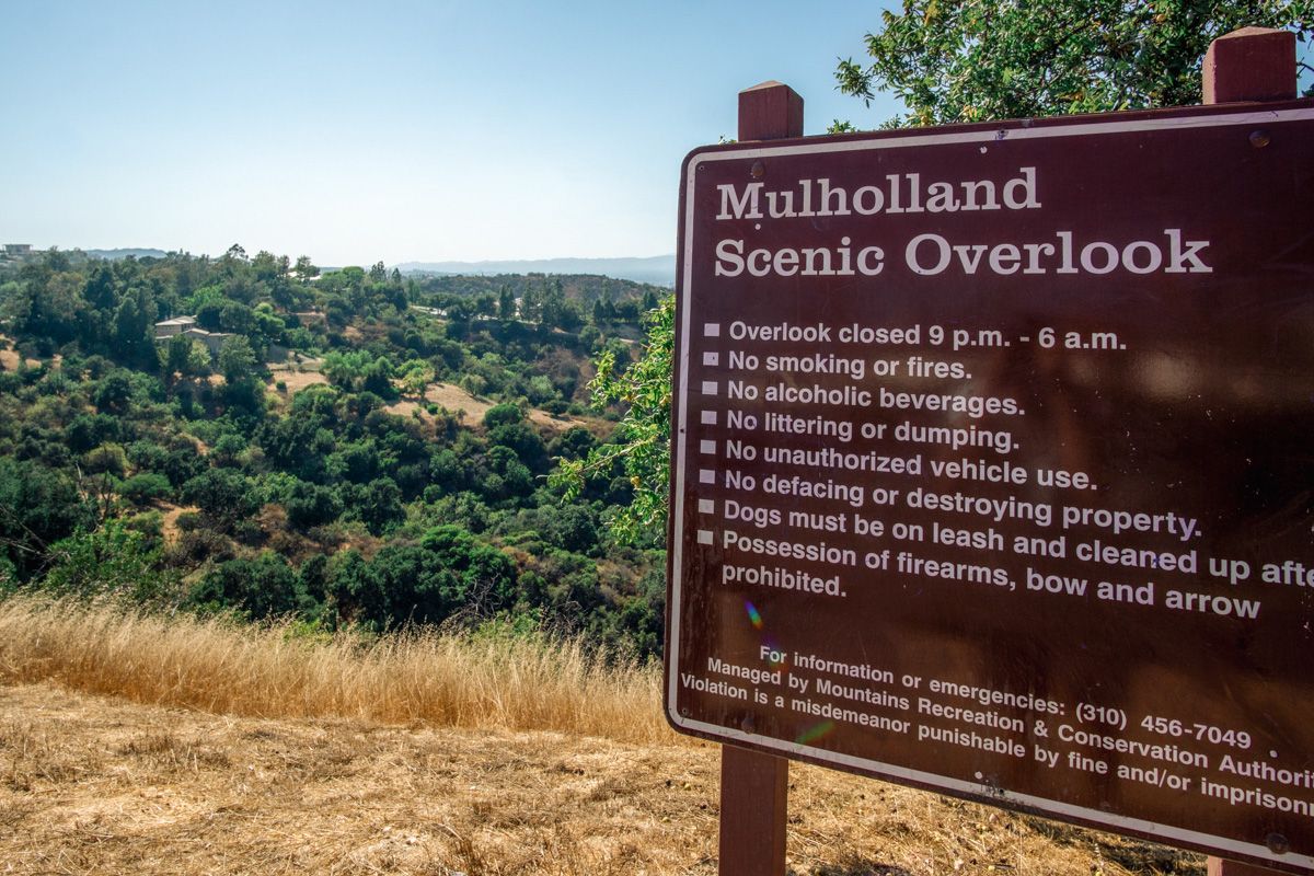 A brown sign with white text listing the rules for the Mulholland Drive Scenic Overlook, with a view of dry golden grass and rolling California Hills beyond.