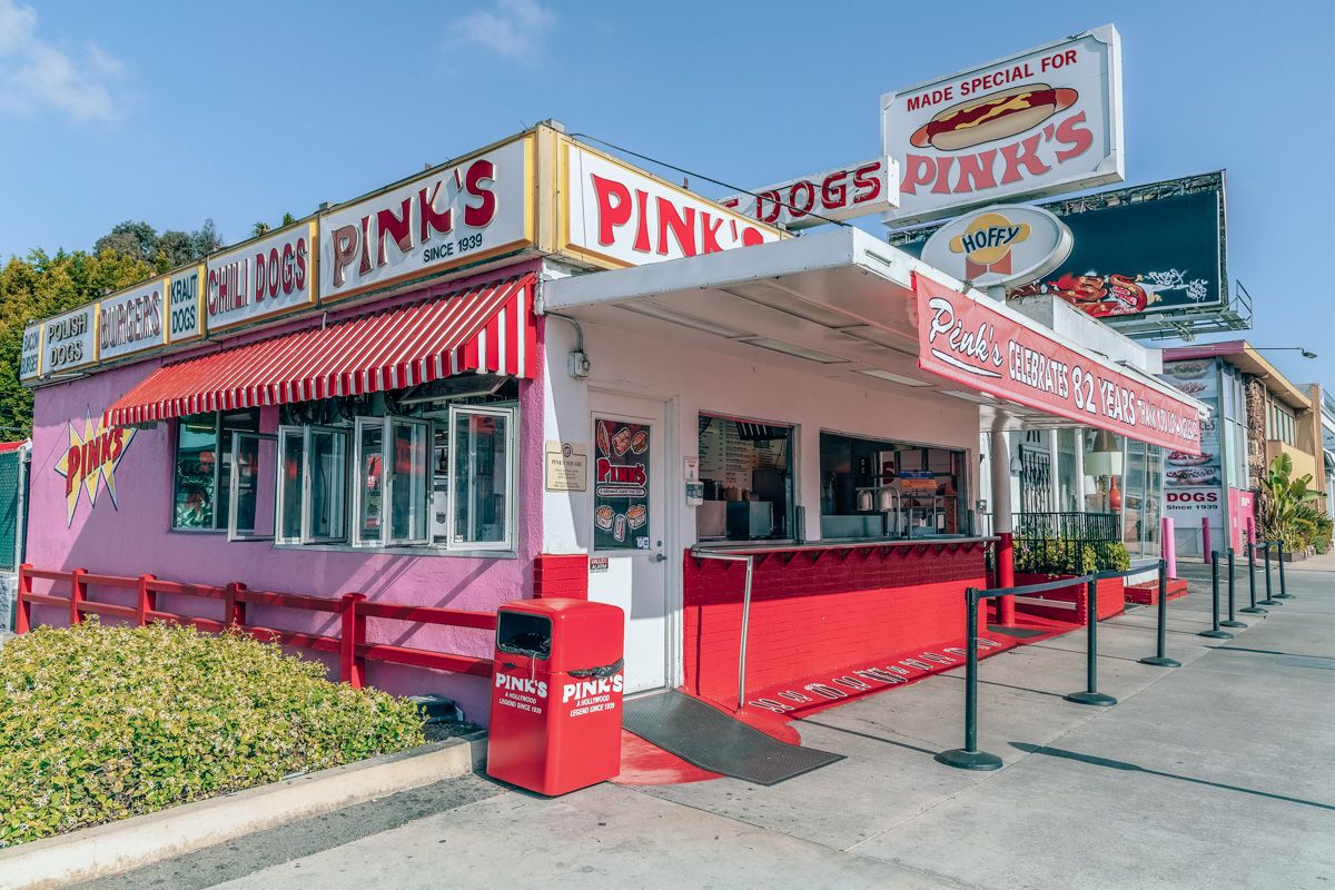 The outer facade of Pink's Hot Dogs stand, a pink building with red and white striped awning, with a clear blue sky beyond.