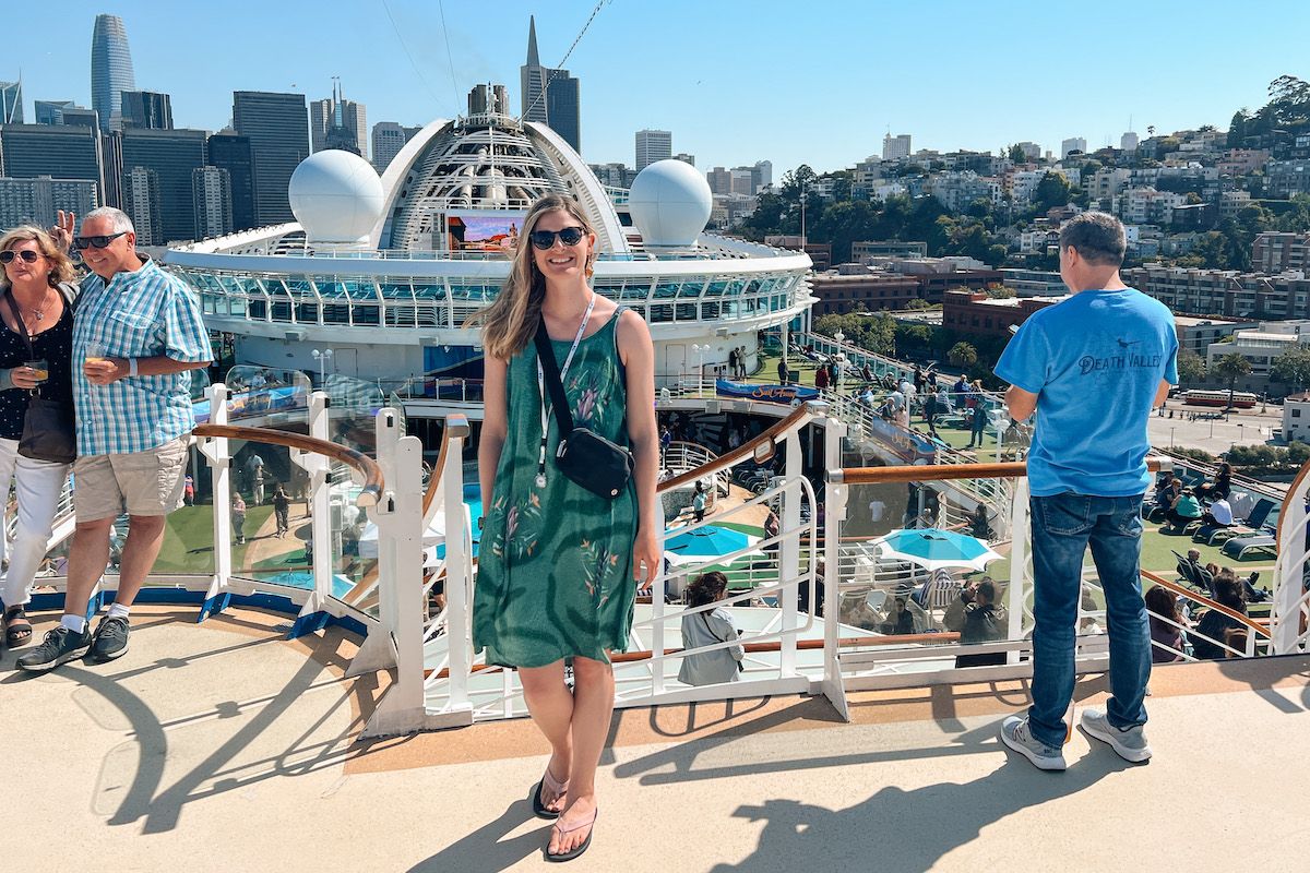 A woman wearing a green printed sundress, sunglasses, and flip flop sandals smiles at the camera from a balcony that overlooks downtown San Francisco.
