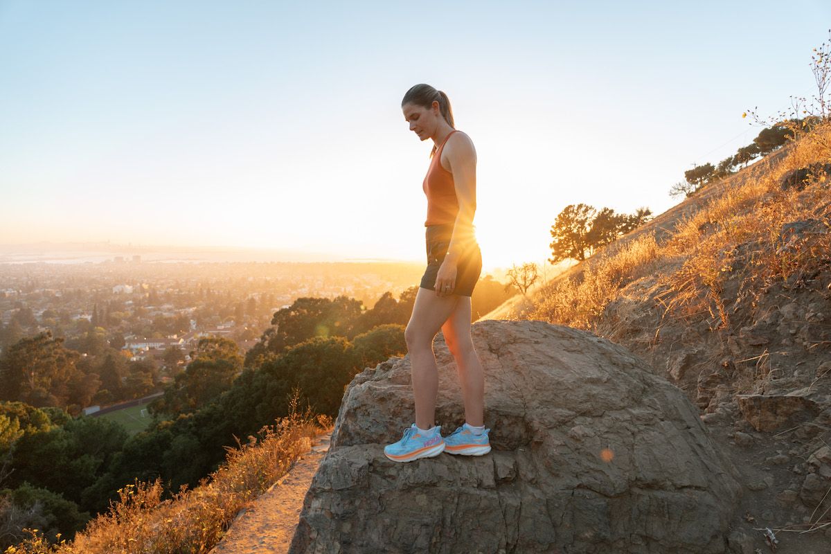 A young woman wearing light blue Hoka walking shoes, an orange tank top, and navy shorts stands on a rock on a grassy hillside overlooking a cityscape, backlit by a golden setting sun. 