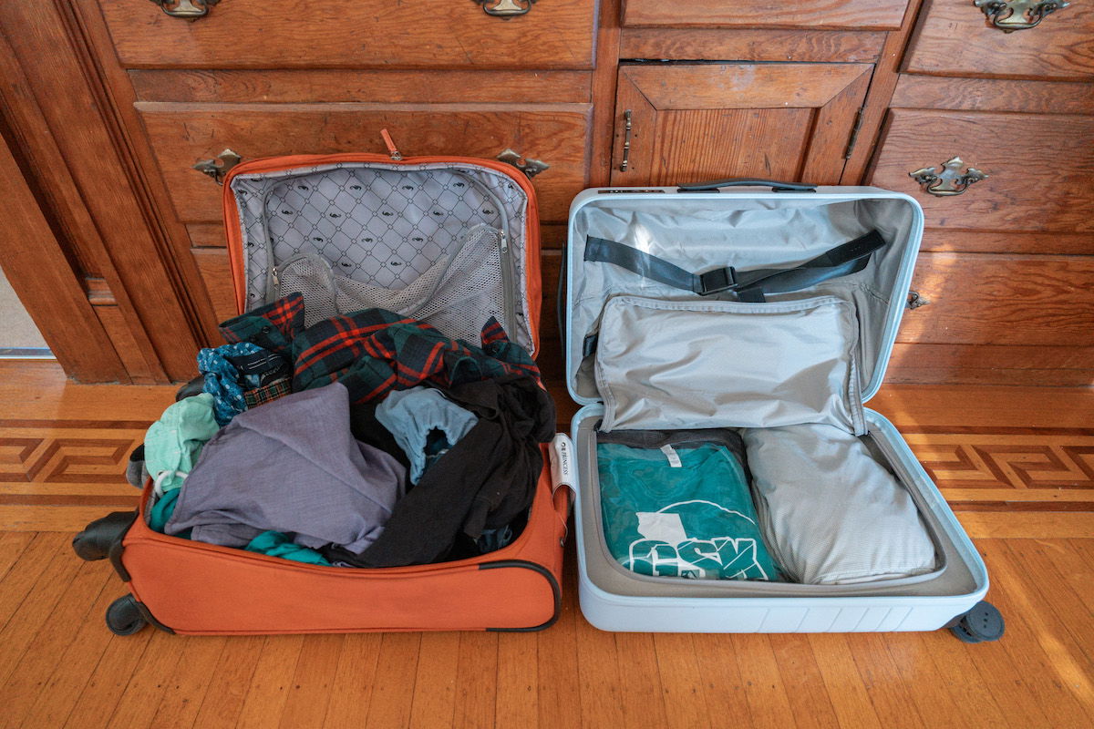 A messily-packed orange suitcase without packing cubes sits open beside a neat, light blue suitcase full of packing cubes on a wooden parquet floor.
