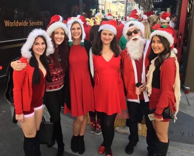 A group of five young women 
 and one young man stand in a lane posing for the picture, all dressed like Santa Claus at the SantaCon Bar Crawl in San Francisco.