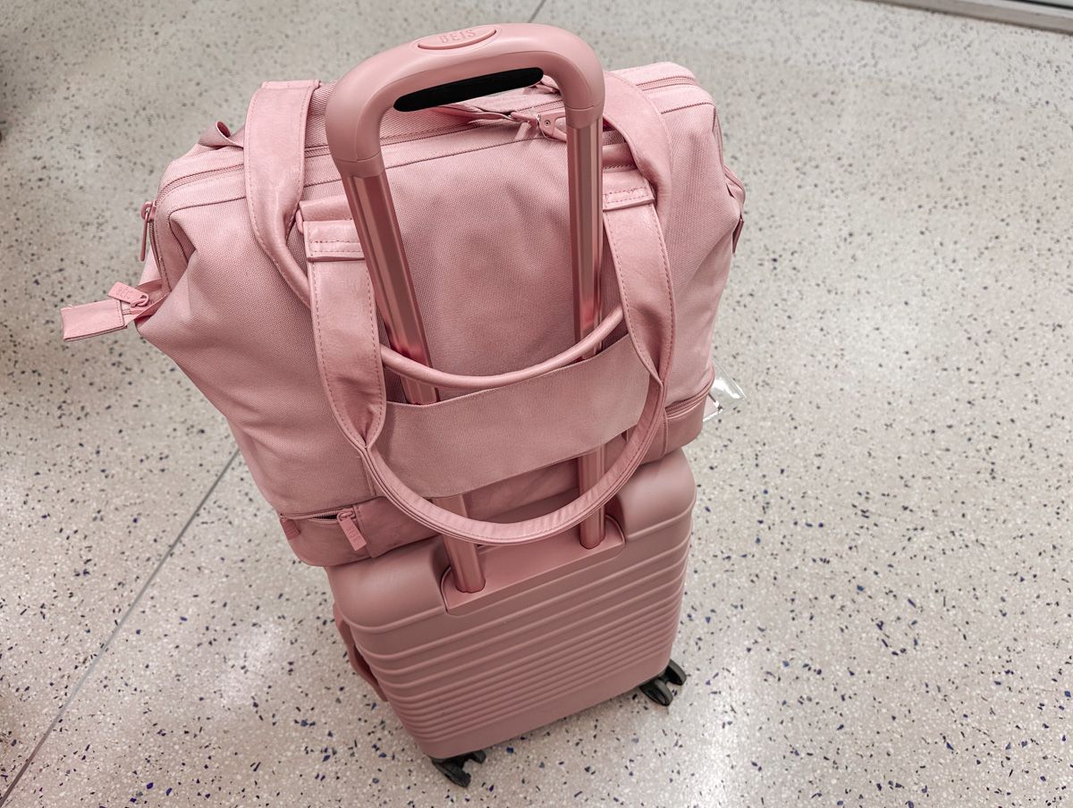 A pink Beis Mini Weekender sitting on top of a pink 21" Béis Luggage Carry On sitting on a grey-flecked airport floor. 