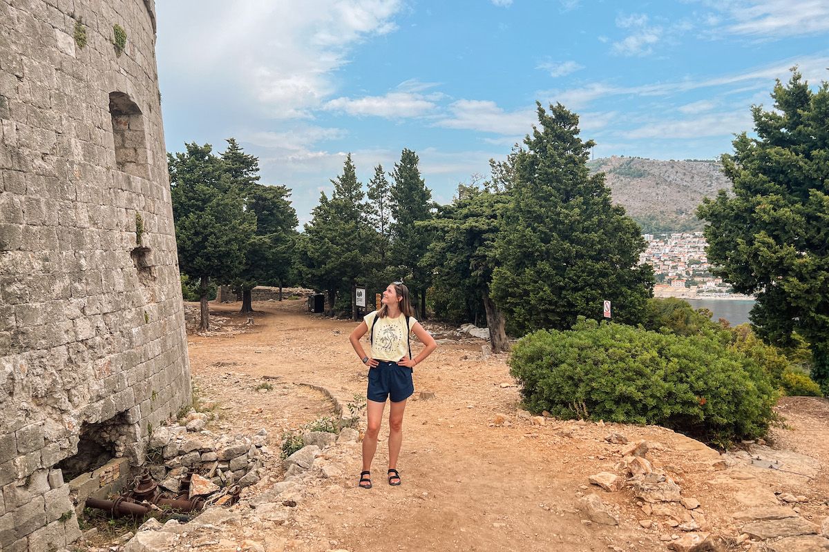 A young woman wearing dark blue sorts, a yellow t-shirt, and black sandals stands with her hands on her hips, looking up at a stone castle wall in a mediterranean landscape.
