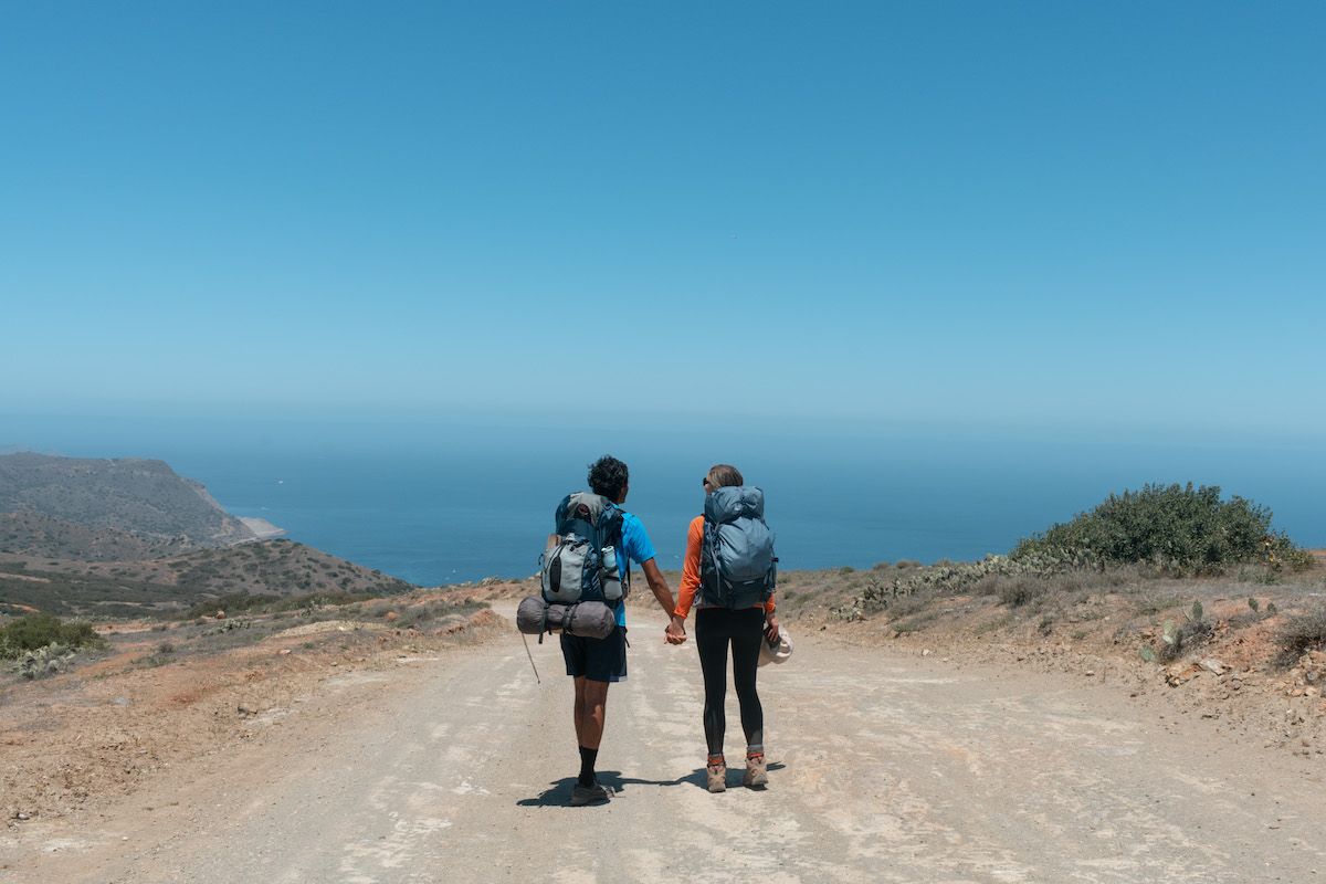 A man and woman wearing backpacking packs hold hands as they hike along a wide trail overlooking the sea, with a clear blue sky beyond it. 