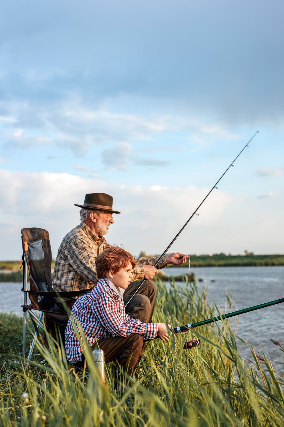 An older man in a brown, brimmed hat holding a fishing pole sits in a chair in tall grass on the banks of a lake with a young, red-haired boy in a checkered shirt sitting beside him holding a fishing pole.