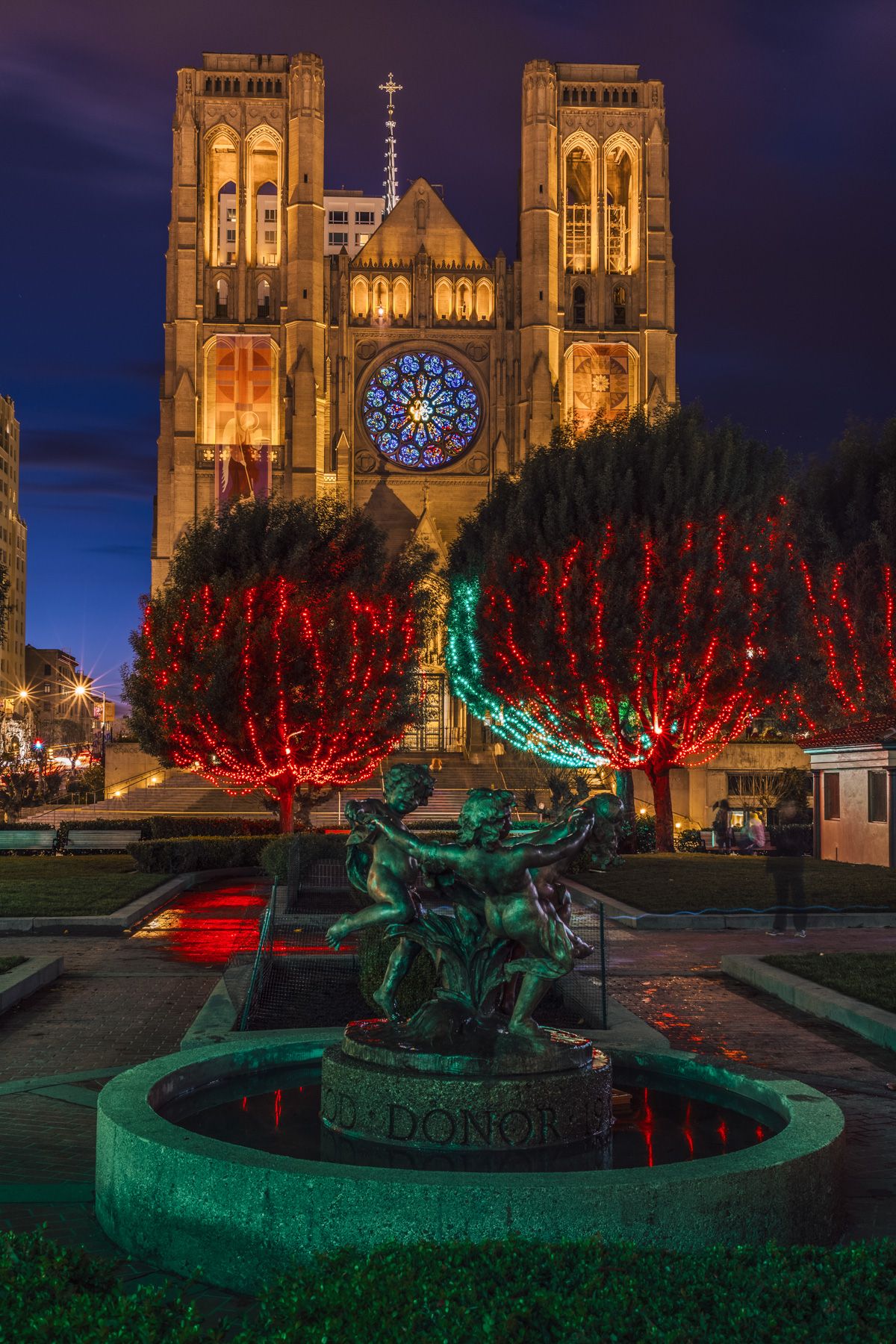 Statues of cherubs playing in the center of a fountain glowing with green light, with rees decorated with red Christmas lights in front of Grace Cathedral in San Francisco, seen at night glowing with yellow light.