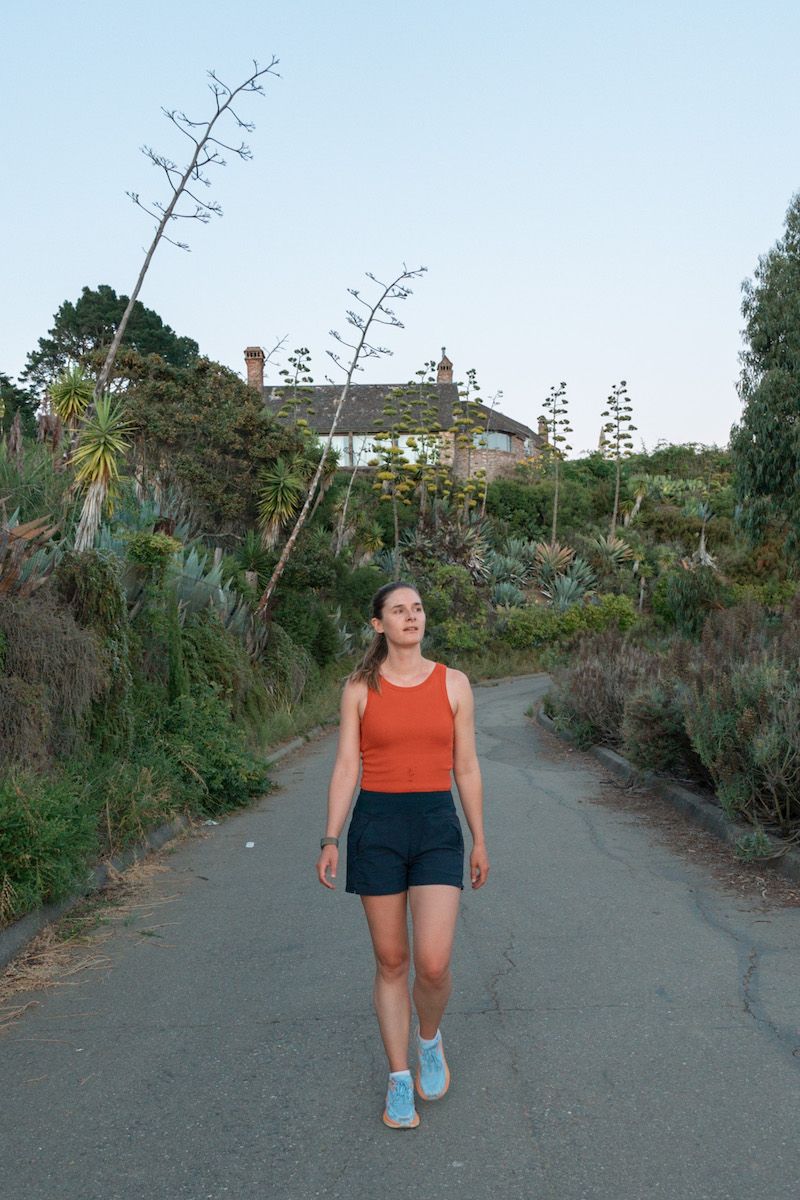 A young woman wearing light blue Hoka walking shoes, an orange tank top, and navy shorts walks towards the camera on a paved path lined by succulents and bushes. 