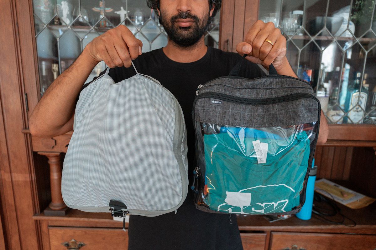 A picture of a man taken at chest level as he holds up two different styles of packing cubes with a wooden cabinet behind him.