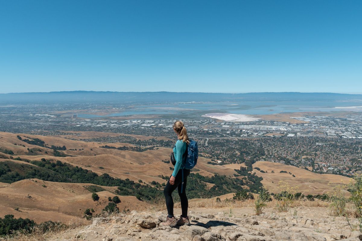 A woman with a ponytail wearing black leggings and a blue top hikes along a ridge that looks out over rolling California hills, and a cityscape and a bay beyond.
