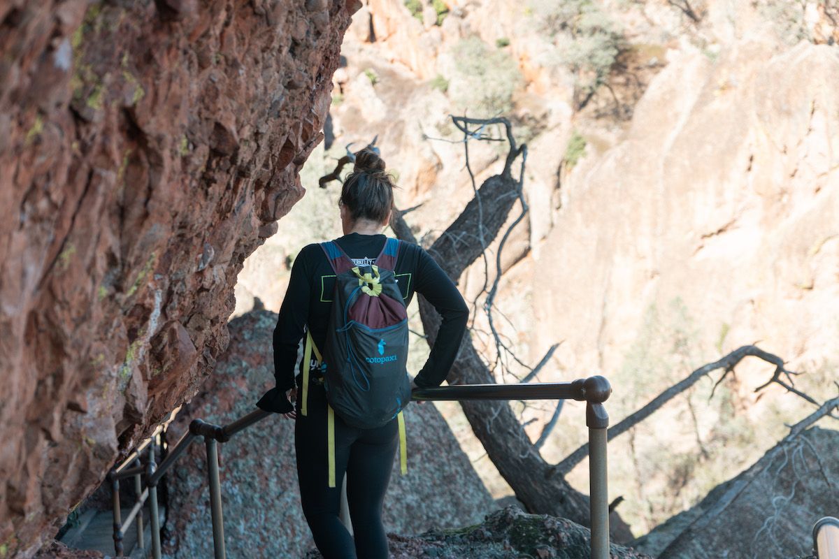 A woman wearing a hiking backpack with brown hair tied up in a bun seen from behind as she hikes down a steep, railed path alongside a red boulder.