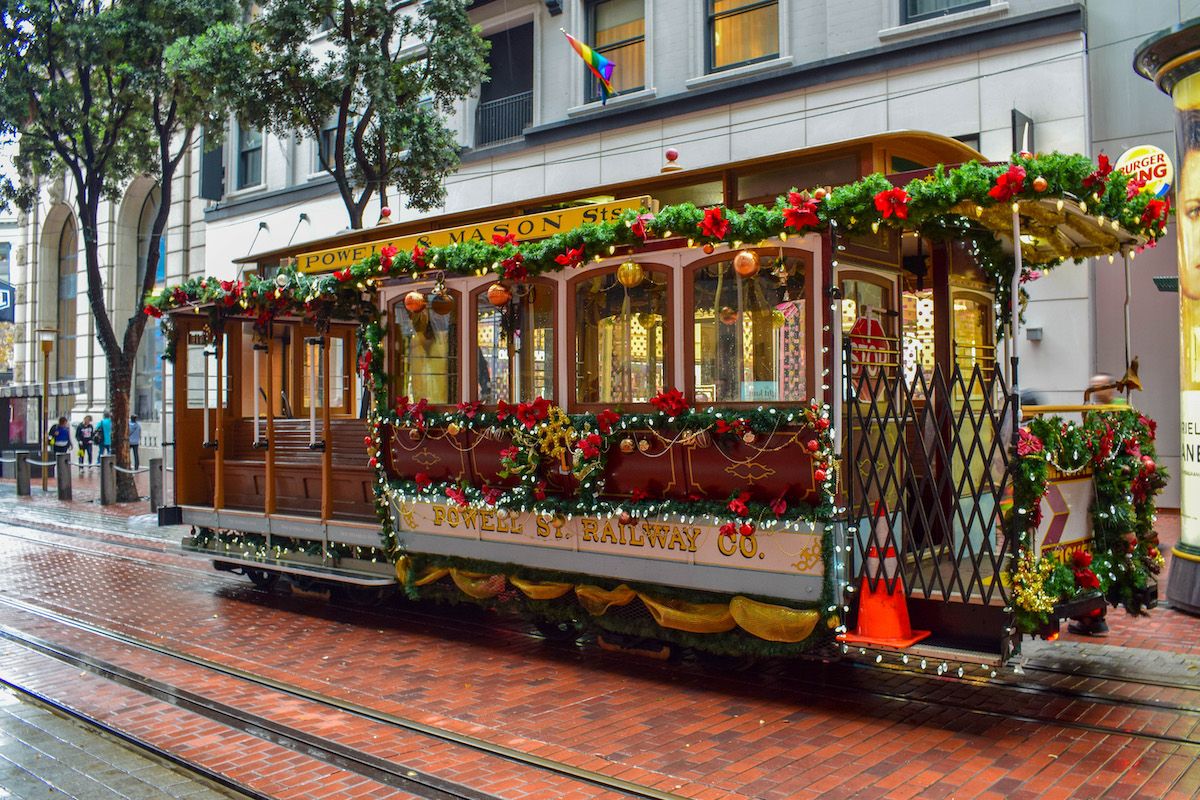 A cable car decorated with wreaths and bows for Christmas in San Francisco, driving over a wet-looking brick street.