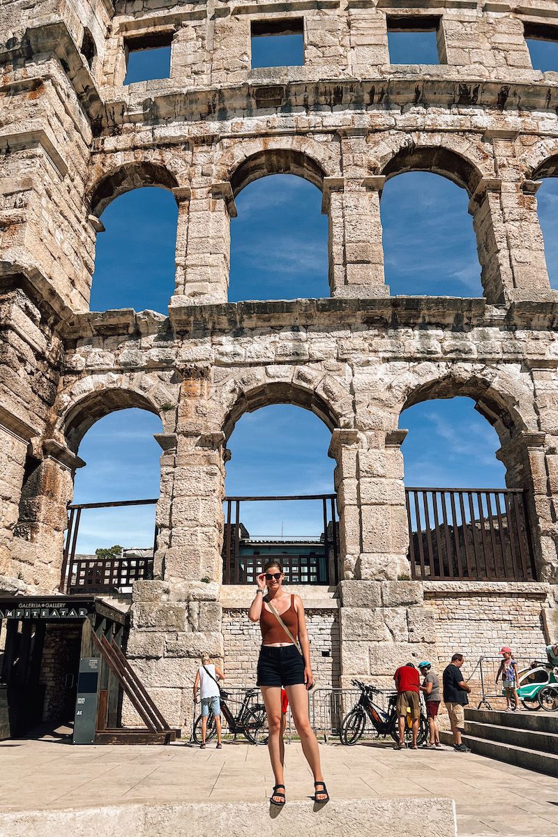 A young woman wearing black shorts, a brown tank top and black Teva sandals stands in front of a Colosseum lookalike in Croatia.
