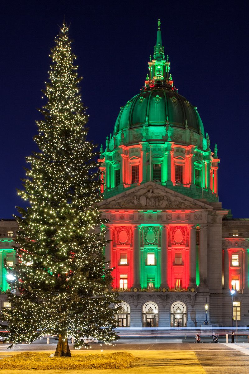 San Francisco City Hall seen at night illuminated by red and green lights, with a large Christmas tree in the foreground.