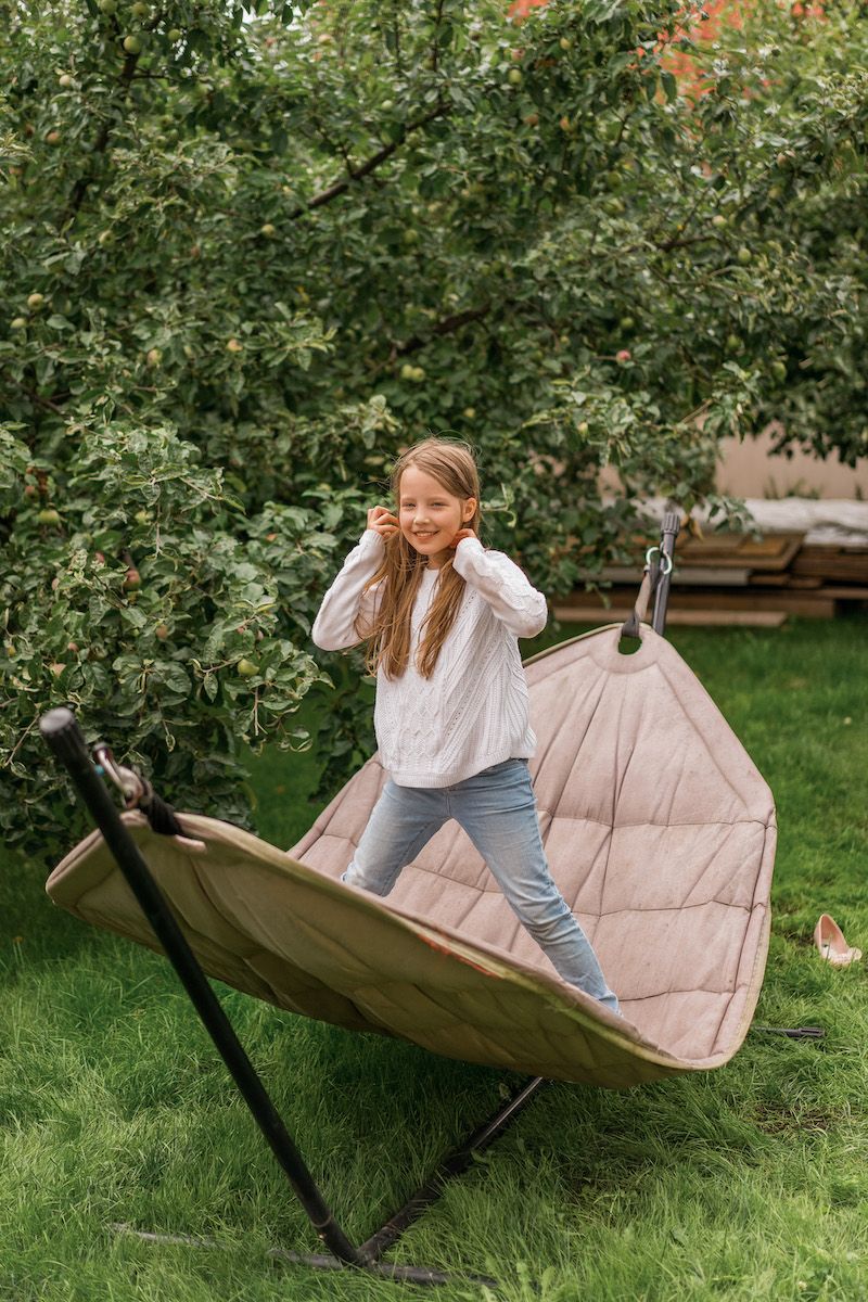 A young blonde girl wearing jeans and a white sweater stands on a grey hammock supported by a stand next to an apple tree in a backyard setting.