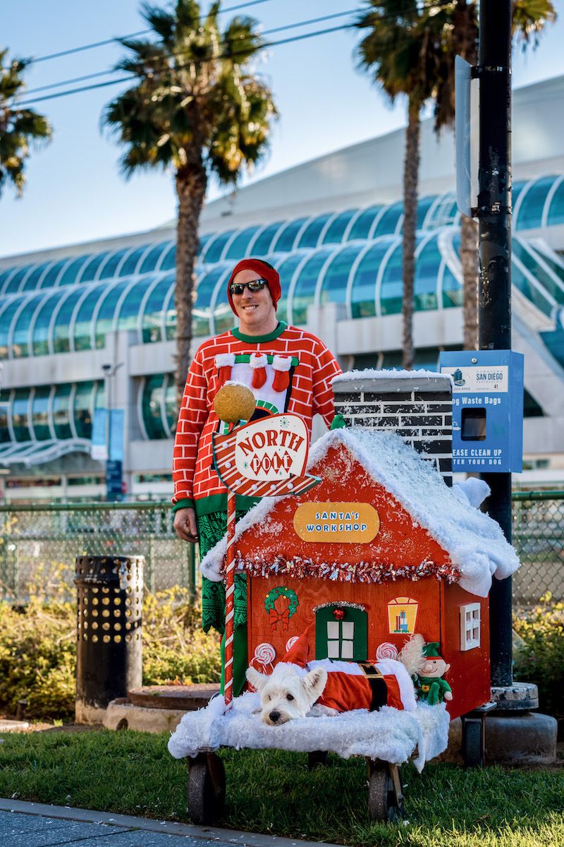 A man wearing an ugly Christmas sweater stands next to a small doghouse decorated like Santa's workshop, with a small, white dog in a Santa costume lying out front, and palm trees in the background.
