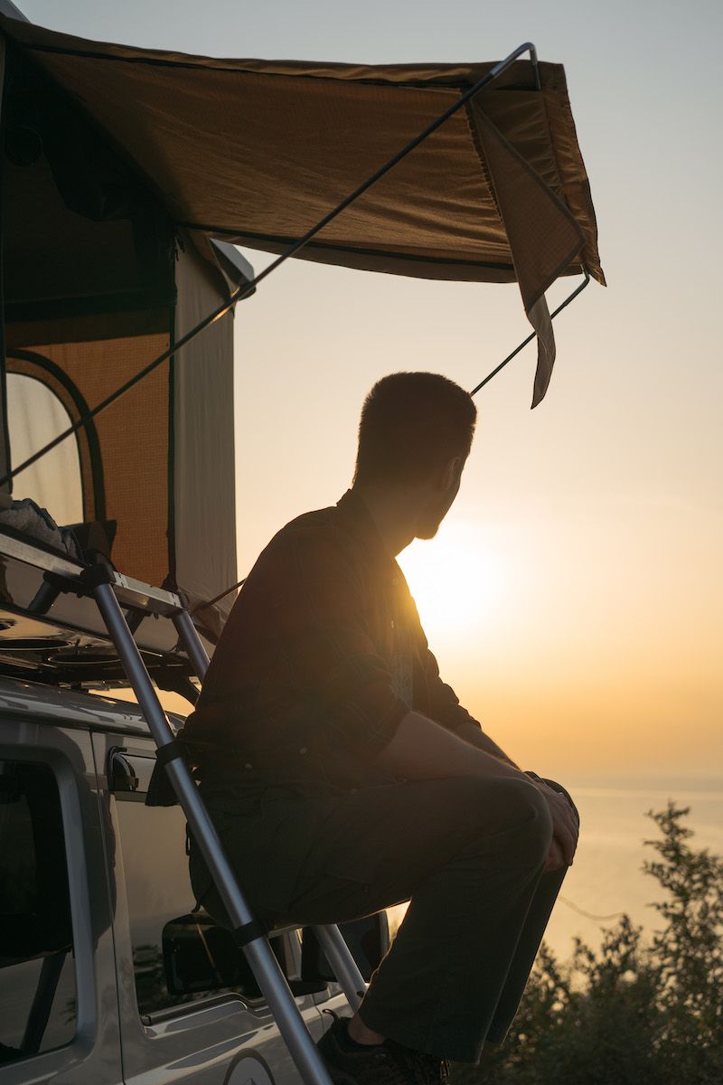The silhouette of a man sitting on the ladder to a truck tent, backlit by a setting sun.
